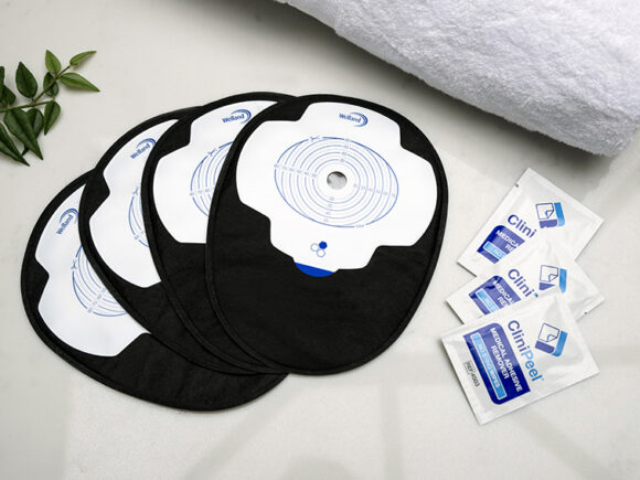 Four black closed Aura Plus stoma bags placed on top of each other and fanned out on a white marble countertop with three sachets of CliniPeel medical adhesive remover beside them.