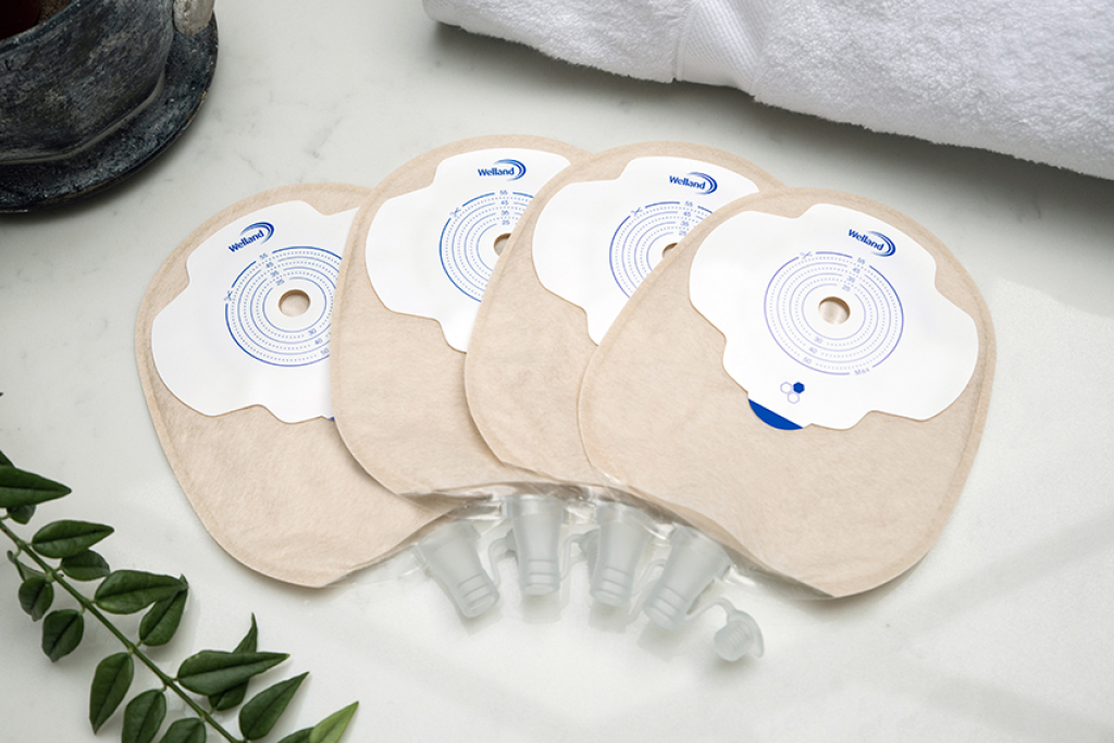 Four sand coloured Aura Plus flat urostomy bags stacked on top of each other and fanned out on a white marble countertop.