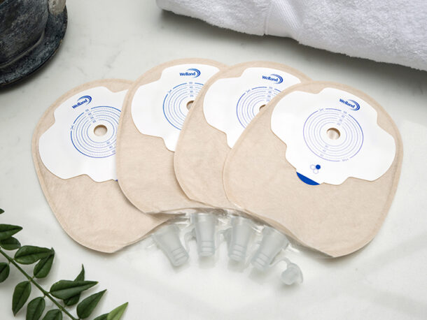 Four sand coloured Aura Plus flat urostomy bags stacked on top of each other and fanned out on a white marble countertop.