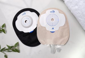 A black closed Aura Plus Soft Convex stoma bag behind a sand coloured flat Aura Plus urostomy bag on a white marble countertop.