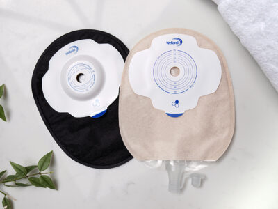 A black closed Aura Plus Soft Convex stoma bag behind a sand coloured flat Aura Plus urostomy bag on a white marble countertop.