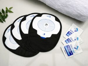 Four black closed Aura Plus stoma bags placed on top of each other and fanned out on a white marble countertop with three sachets of CliniPeel medical adhesive remover beside them.