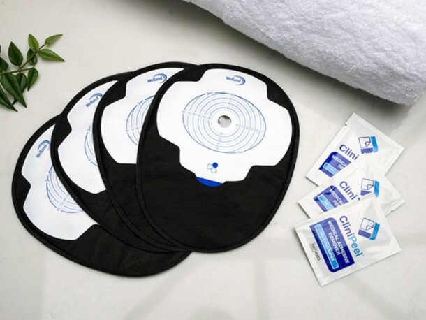 Four black closed Aura Plus stoma bags placed on top of each other and fanned out on a white marble countertop with three sachets of CliniPeel medical adhesive remover beside them.