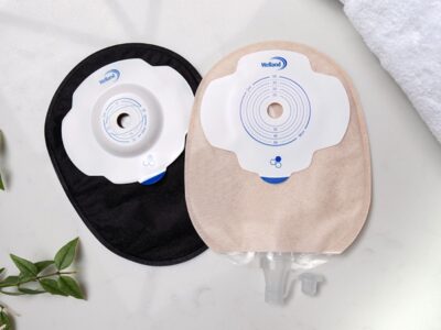 A black Aura Plus Soft Convex closed bag lies under a sand Aura Plus urostomy bag on a marble countertop