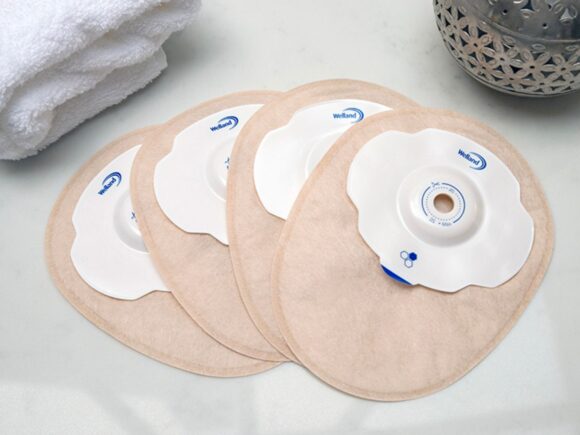 Four sand coloured closed Aura Plus Soft Convex stoma bags placed on top of each other and fanned out on a white marble countertop.