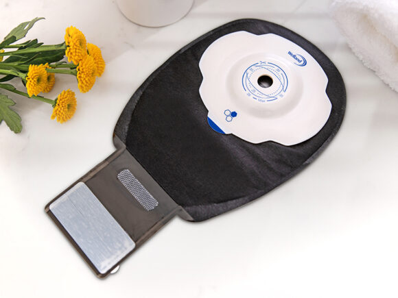 A black Aura Plus Soft Convex drainable ileostomy bag with the opening unfurled on a white marble worktop.
