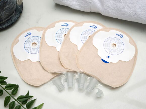 Four sand coloured Aura Plus flat urostomy bags stacked on top of each other and fanned out on a white marble countertop.