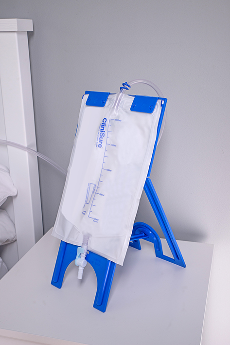 A CliniSure sterile night drainage bag on a blue plastic stand on a bedside table.