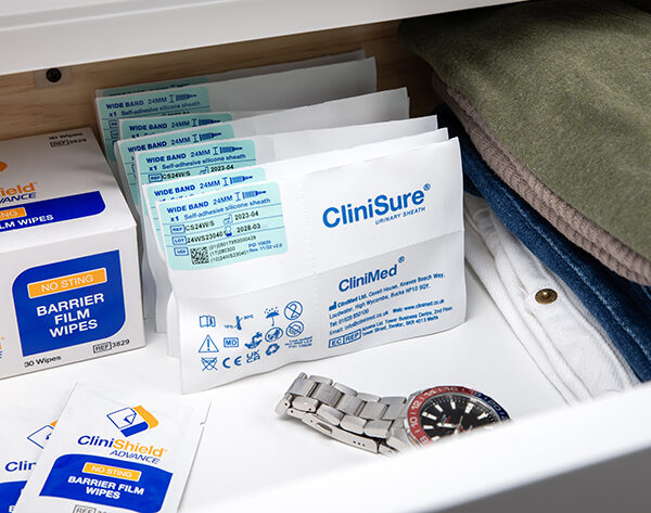 Seven CliniSure sheaths in their packaging with three CliniShield Advance wipe sachets and a box of wipes in a drawer with folded clothing and a watch.