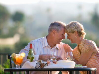 An older couple sit at a table on a terrace, the sun is low and they are enjoying a meal and wine. They lean in towards one another and touch foreheads while smiling and laughing.