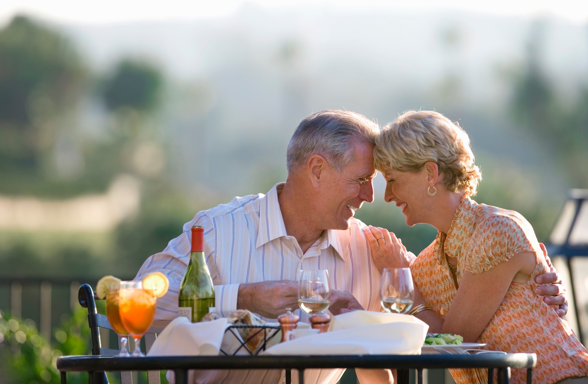 An older couple sit at a table on a terrace, the sun is low and they are enjoying a meal and wine. They lean in towards one another and touch foreheads while smiling and laughing.