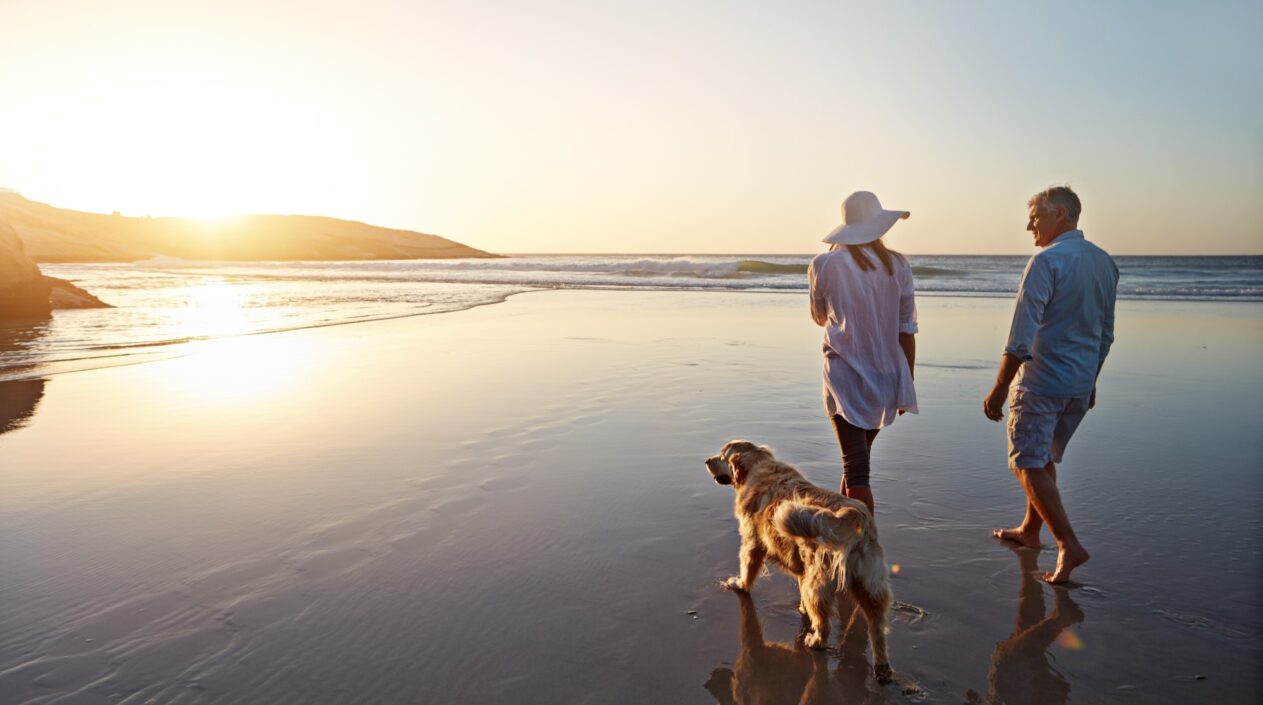 A middle-aged couple and their golden retriever walk on a beach as the tide comes in. The sun is setting.