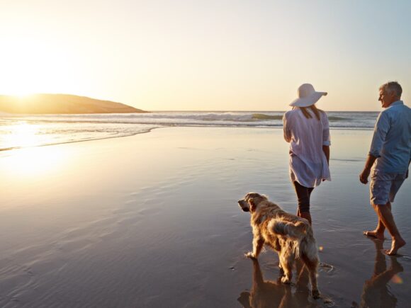 A middle-aged couple and their golden retriever walk on a beach as the tide comes in. The sun is setting.