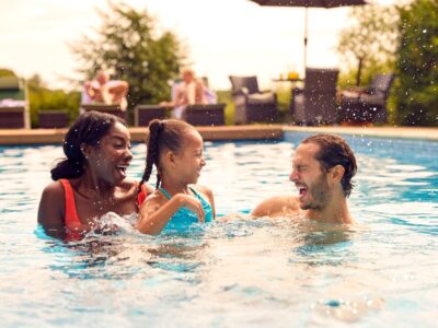 A couple play in an outdoor swimming pool with their child, the little girl splashes her father, they are all smiling.