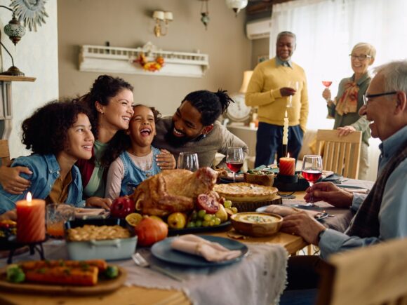 A family sits at a dining table laid with a roast dinner, the smiling parents hug their children while the grandparents look on and laugh.