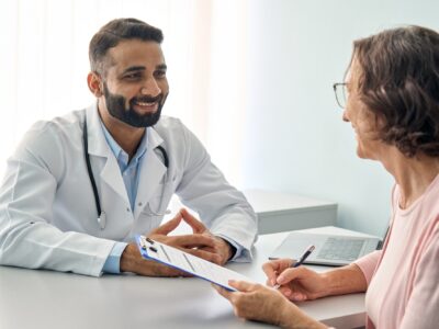 A young GP with a well-trimmed beard sits across from an elderly patient who is filling out paperwork attached to a clipboard, they are smiling at each other.