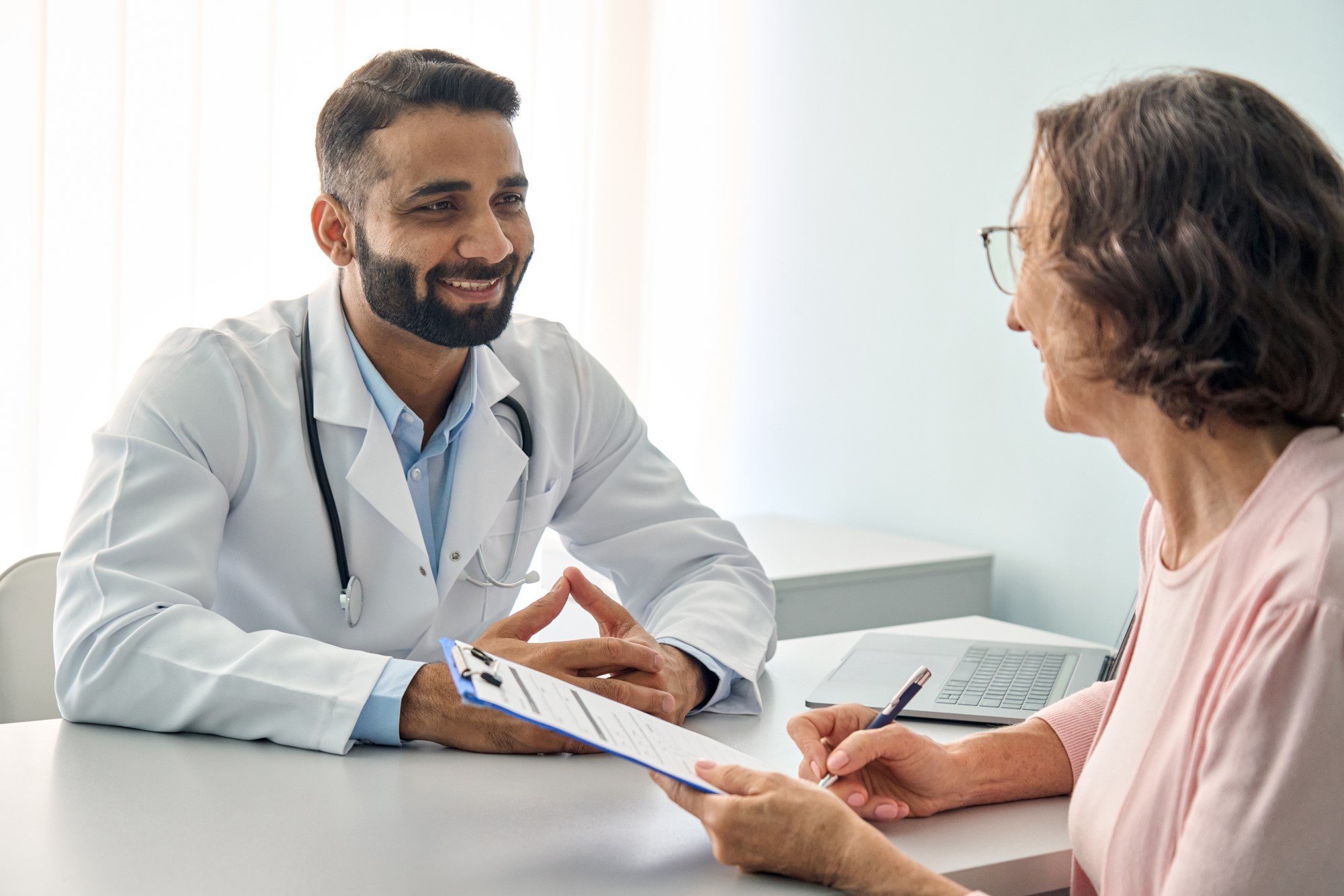 A young GP with a well-trimmed beard sits across from an elderly patient who is filling out paperwork attached to a clipboard, they are smiling at each other.