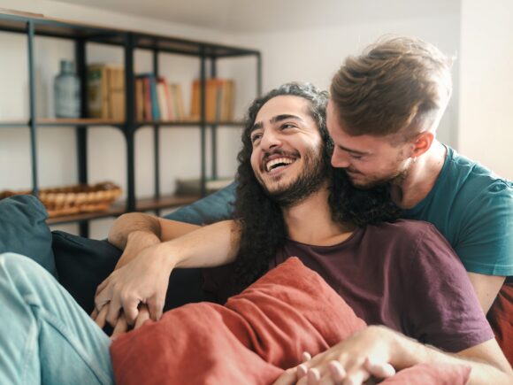 A gay couple sit on a sofa, one man sits with his back against his partner who looks over his shoulder as they both laugh. They interlink their fingers on both hands.