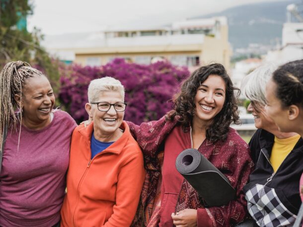 A group of women of different ages talk and laugh outside in a city while wearing exercise clothes and carrying yoga mats.