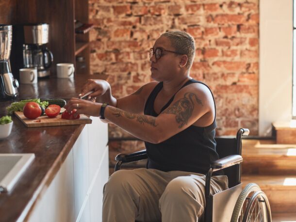 A woman with short hair and tattoos wearing glasses sits in her wheelchair next to a kitchen worktop and cuts vegetables.