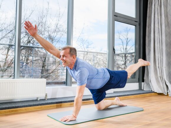 A man kneels on a yoga mat in a room with hardwood floors and tall windows, he is performing yoga exercise and smiling.