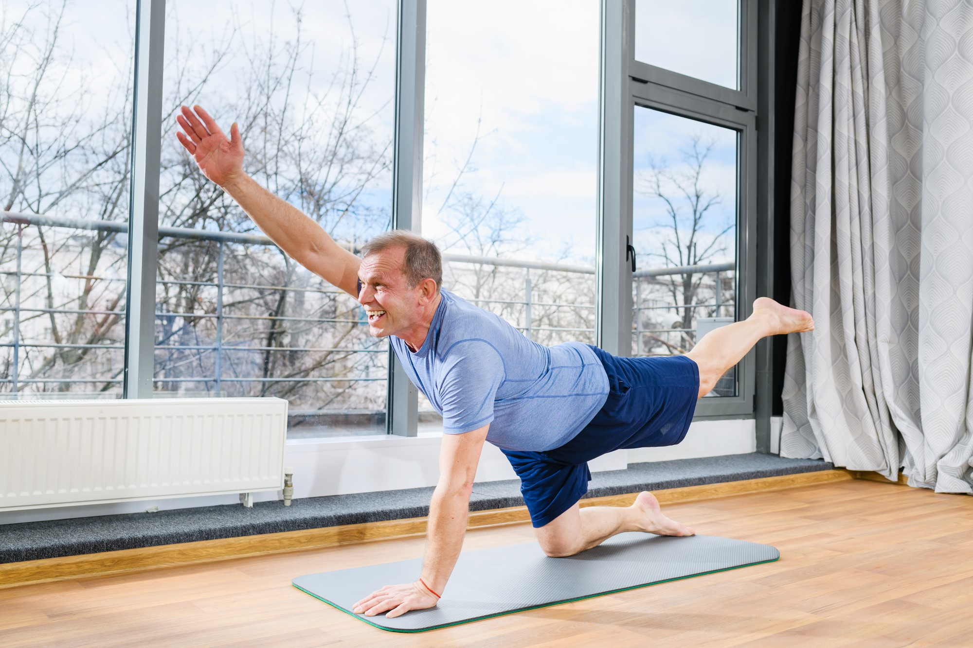 A man kneels on a yoga mat in a room with hardwood floors and tall windows, he is performing yoga exercise and smiling.