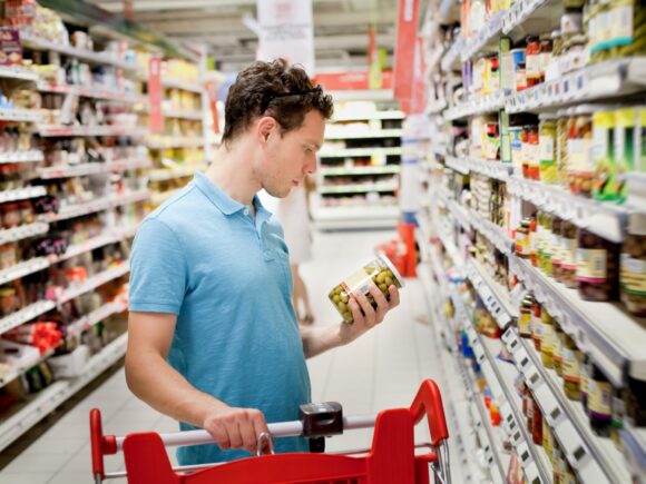 A man stands in a supermarket aisle one hand resting on his trolley in the other he holds a jar of olives, his attention is fully absorbed reading the label on the jar.