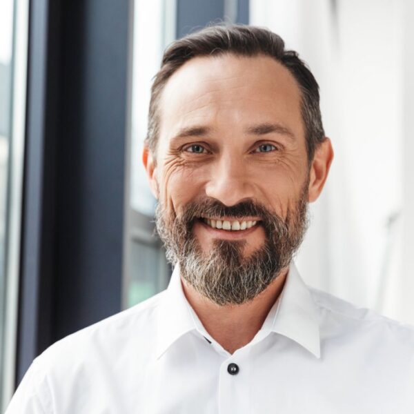 A man with short brown hair and a mid-length, greying brown beard smiles at the camera, the skin around his blue eyes crinkles.