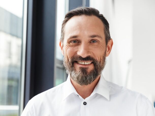 A man with short brown hair and a mid-length, greying brown beard smiles at the camera, the skin around his blue eyes crinkles.