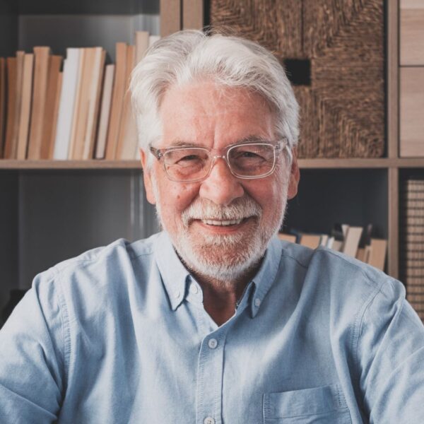 An older man with white hair and a short, white beard sits in front of bookshelves smiling, he wears thick framed glasses and a pale blue button-down shirt.
