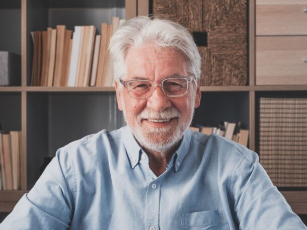 An older man with white hair and a short, white beard sits in front of bookshelves smiling, he wears thick framed glasses and a pale blue button-down shirt.