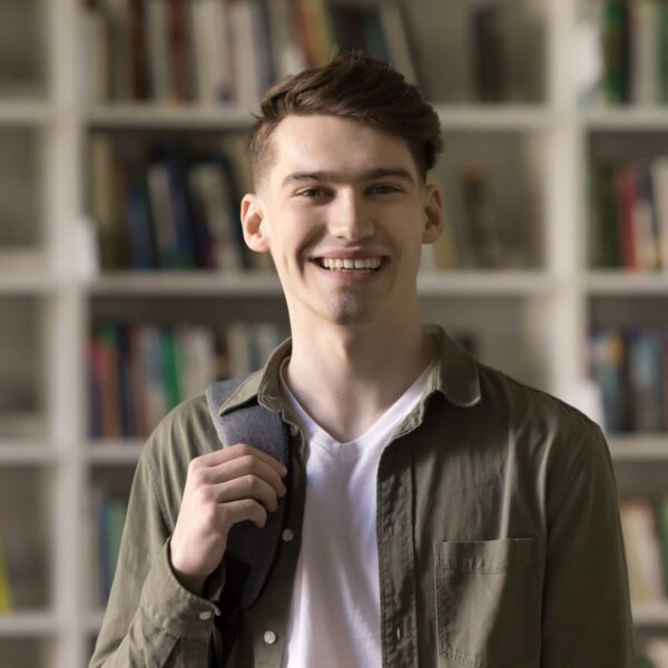 A young adult man with short brown hair stands in front of bookcases with a backpack slung over one shoulder, he smiles broadly at the camera.