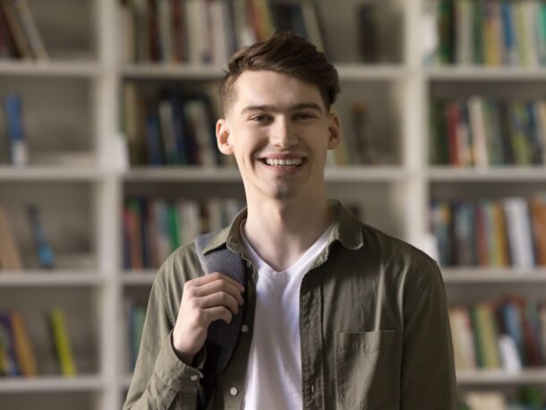 A young adult man with short brown hair stands in front of bookcases with a backpack slung over one shoulder, he smiles broadly at the camera.