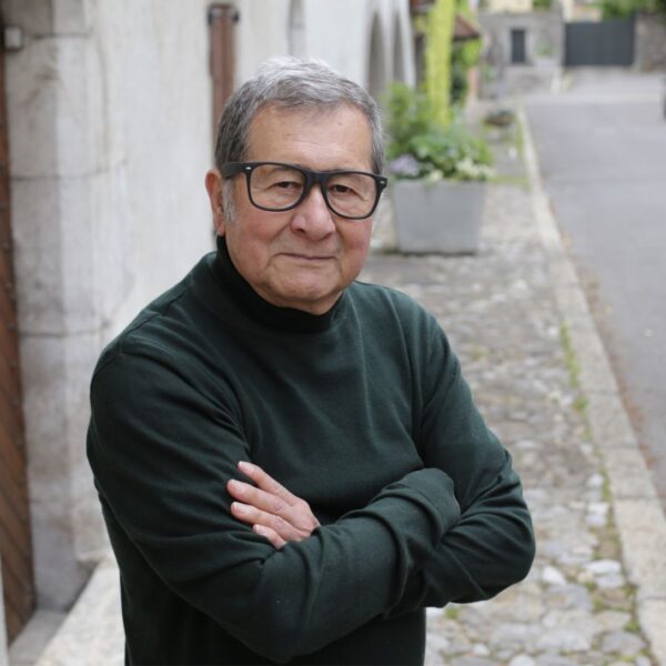 An older man with short greying hair wearing thick rimmed glasses and a black polo neck, stands in the street smiling subtly with his arms crossed.