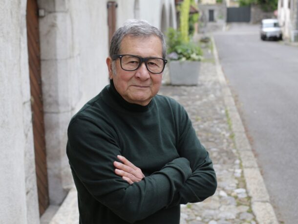 An older man with short greying hair wearing thick rimmed glasses and a black polo neck, stands in the street smiling subtly with his arms crossed.