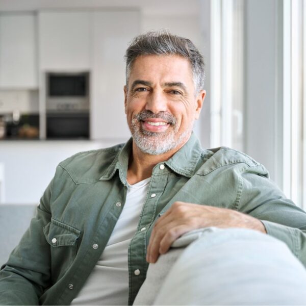 An older man sits sideways-on on a sofa with his arm resting on the back smiling at the camera. He has salt and pepper hair and a short greying beard.