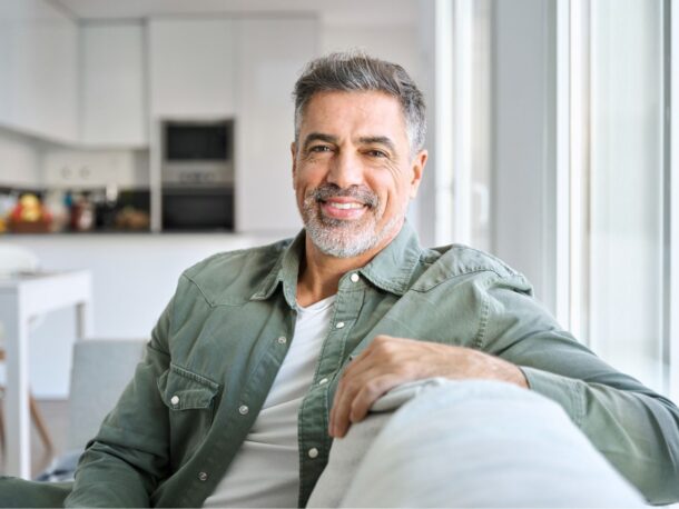 An older man sits sideways-on on a sofa with his arm resting on the back smiling at the camera. He has salt and pepper hair and a short greying beard.