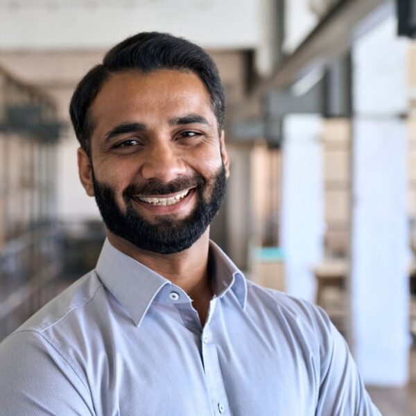 A young man with short black hair and a beard smiles at the camera, he is in an office and wearing a light blue button-down shirt.