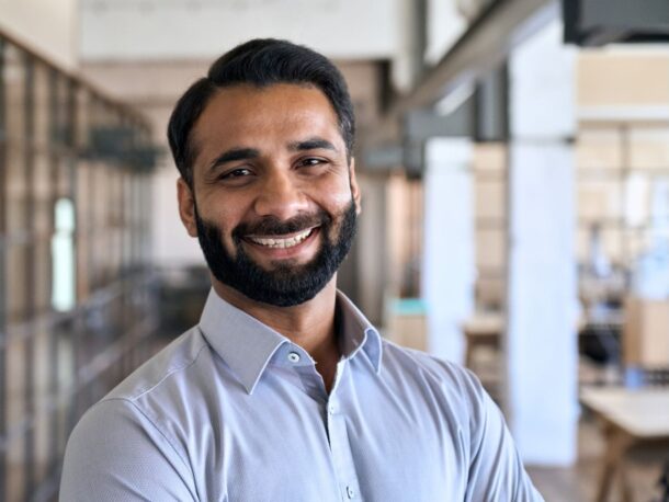 A young man with short black hair and a beard smiles at the camera, he is in an office and wearing a light blue button-down shirt.