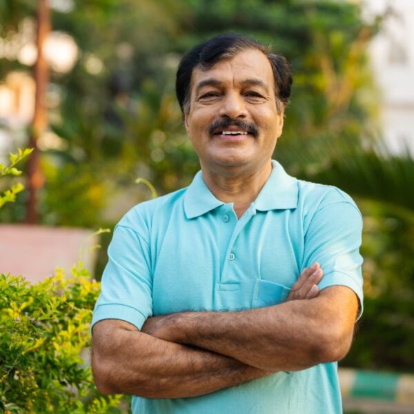 An older man with black hair and a moustache stands in front of a hedge with his arms folded as he smiles.