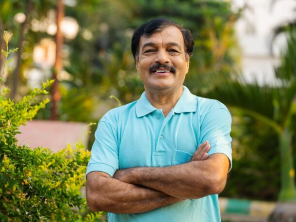 An older man with black hair and a moustache stands in front of a hedge with his arms folded as he smiles.