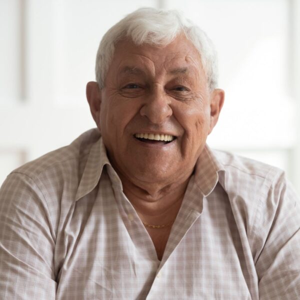 An elderly man with short white hair smiles at the camera as if mid-laugh. He has olive skin and is on the larger side. He's wearing a beige and light brown checked shirt with a few buttons open revealing a gold chain around his neck.