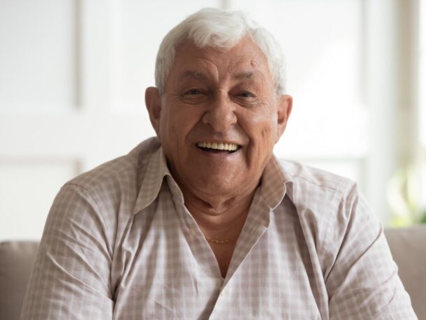 An elderly man with short white hair smiles at the camera as if mid-laugh. He has olive skin and is on the larger side. He's wearing a beige and light brown checked shirt with a few buttons open revealing a gold chain around his neck.