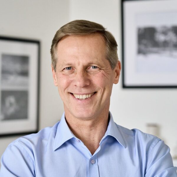 A middle aged man with greying light brown hair, blue eyes and pale freckled skin smiles broadly at the camera. Smile lines wrinkle his eyes. He's wearing a light blue button-up shirt.