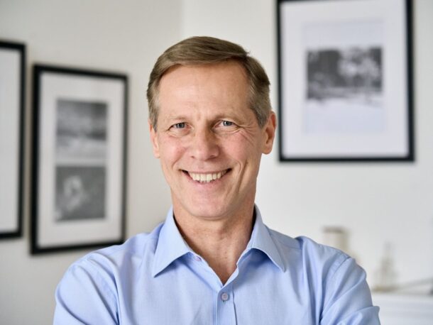 A middle aged man with greying light brown hair, blue eyes and pale freckled skin smiles broadly at the camera. Smile lines wrinkle his eyes. He's wearing a light blue button-up shirt.
