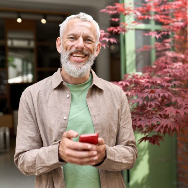 A middle-aged man with, tanned skin, short grey hair and a well-kept beard smiles broadly, lines forming round his blue eyes. He's wearing an open brown button-up shirt with a green t-shirt underneath while holding a red smartphone.