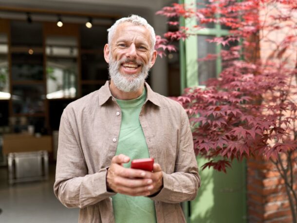 A middle-aged man with, tanned skin, short grey hair and a well-kept beard smiles broadly, lines forming round his blue eyes. He's wearing an open brown button-up shirt with a green t-shirt underneath while holding a red smartphone.