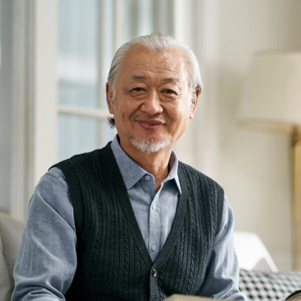 An older man with swept-back white hair and a short white goatee style beard smiles at the camera as he sits on the sofa with an open book in his hands.