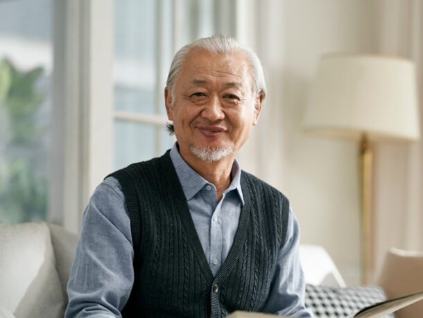 An older man with swept-back white hair and a short white goatee style beard smiles at the camera as he sits on the sofa with an open book in his hands.