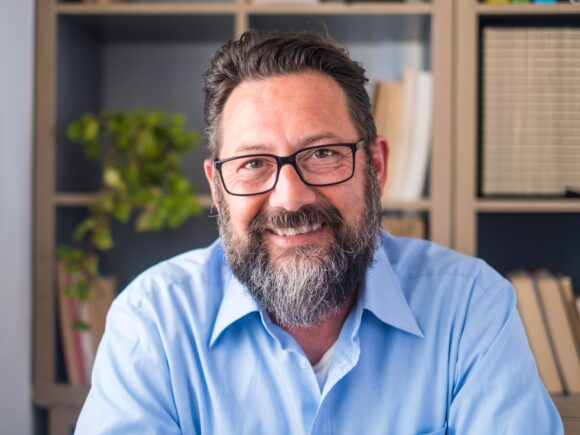 A man with short brown hair and a mid-length greying brown beard smiles at the camera, he wears thick rimmed glasses and a blue button-down shirt.